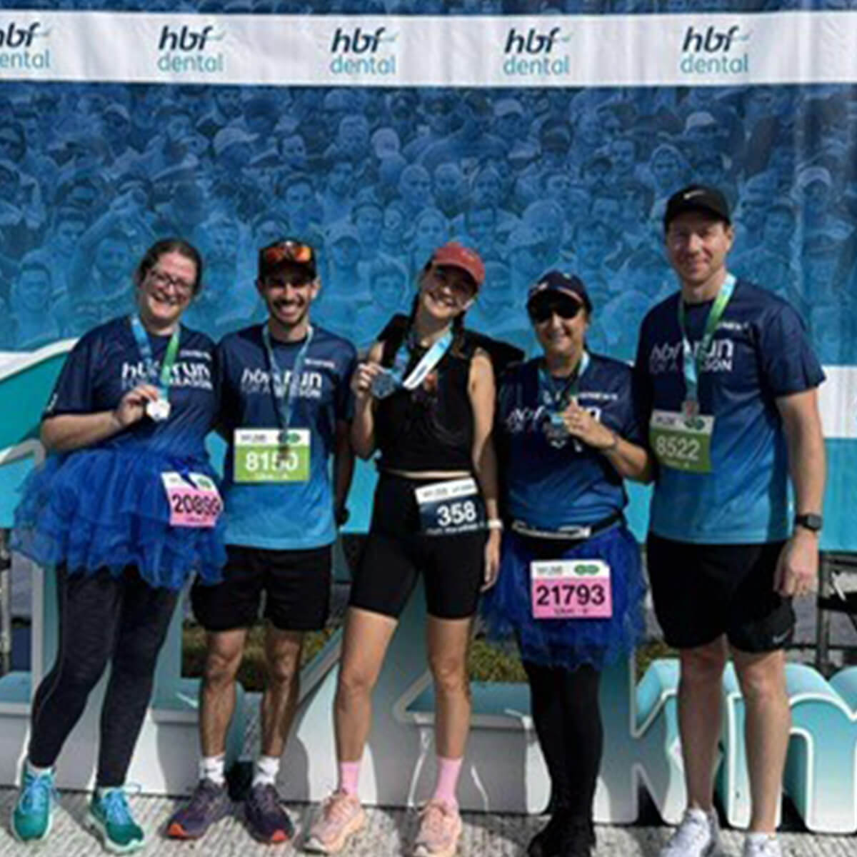 Five participants in athletic gear pose with medals in front of a blue backdrop after a race event.