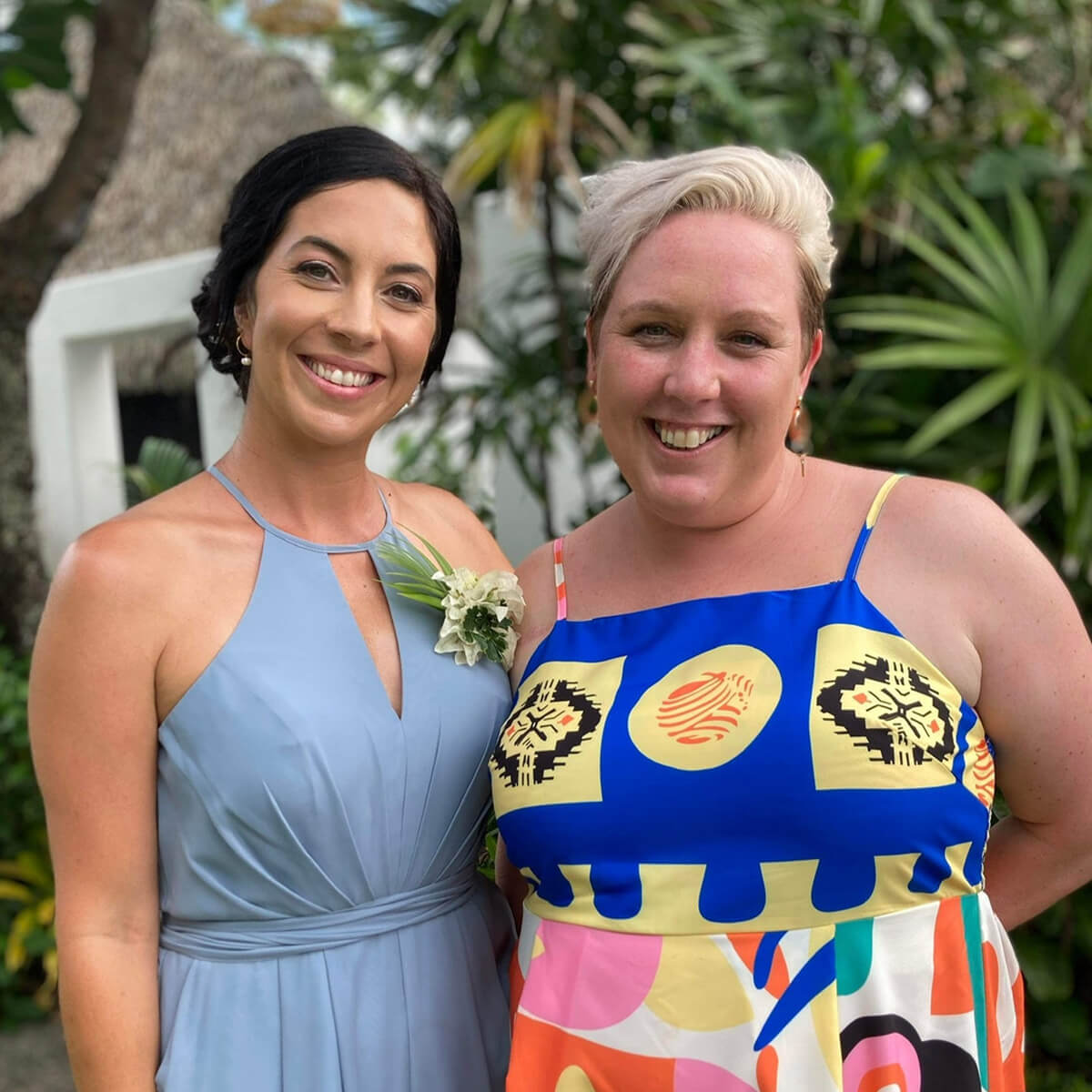 Two women smile for the camera, surrounded by lush greenery. One wears a blue dress with a flower, the other a colorful patterned dress.