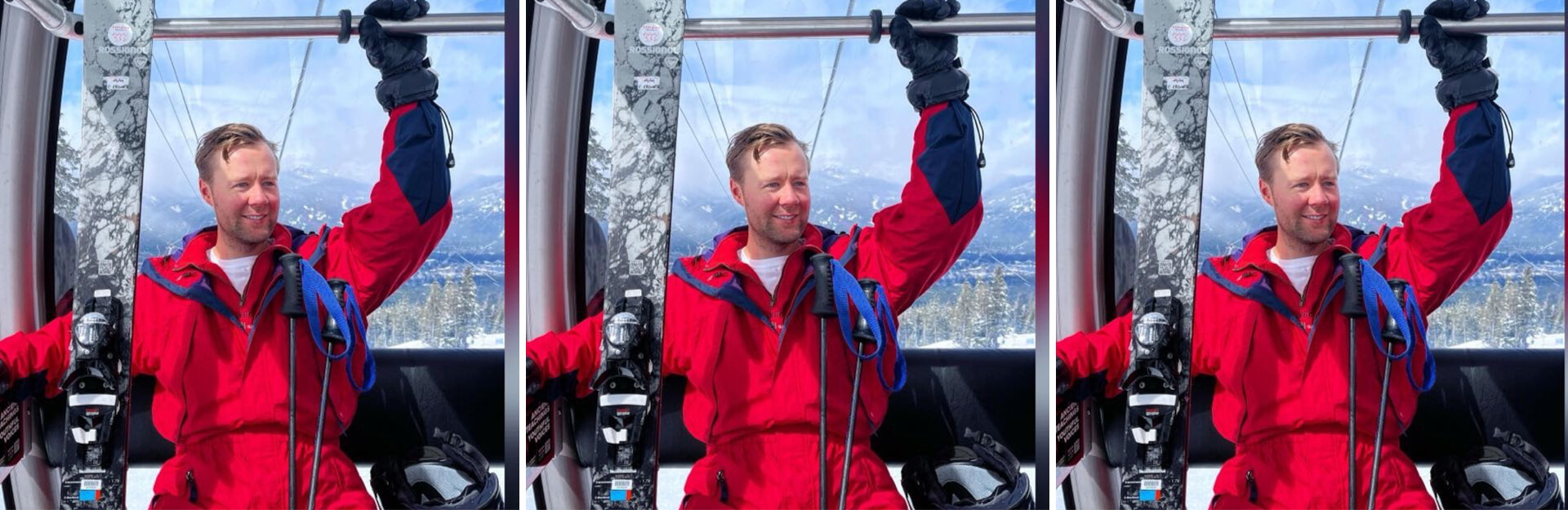 A man in a red ski outfit smiles while holding onto a ski pole in a gondola with snowy mountains in the background.