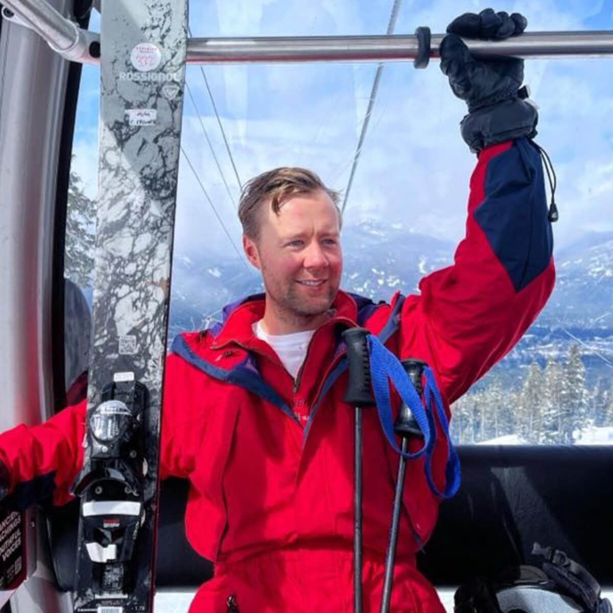 A man in a red ski outfit smiles while holding onto a pole in a ski gondola, with snowy mountains visible outside.