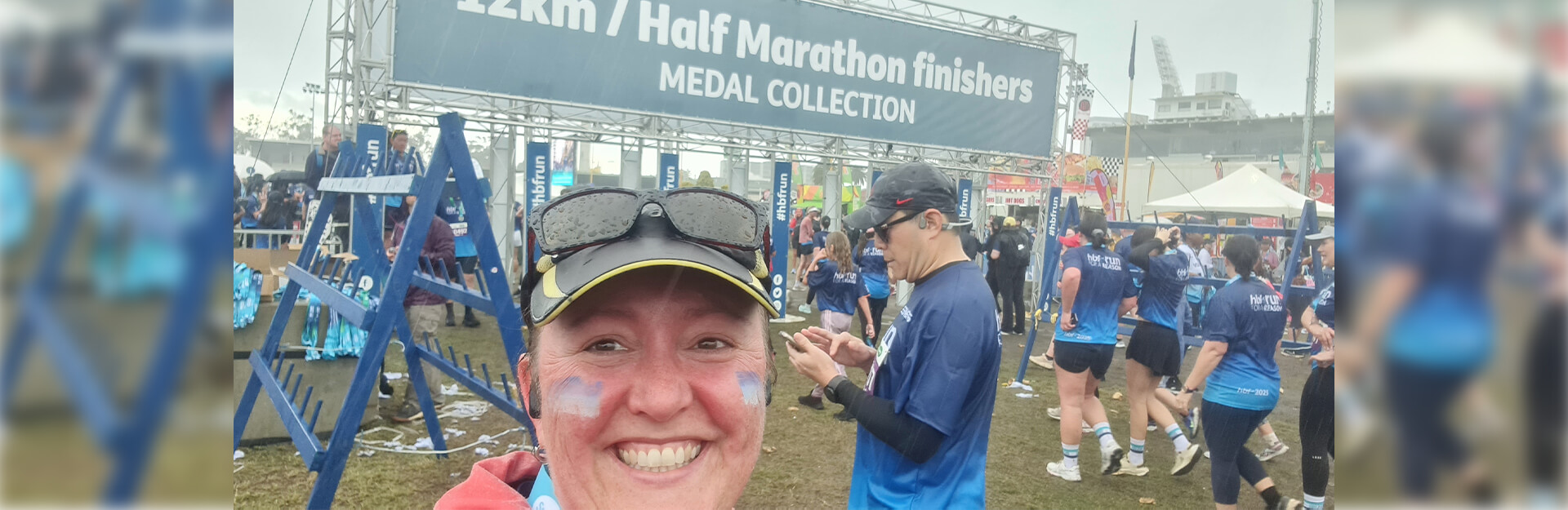 A person takes a selfie at a marathon finish line with a "12km / Half Marathon finishers Medal Collection" sign.