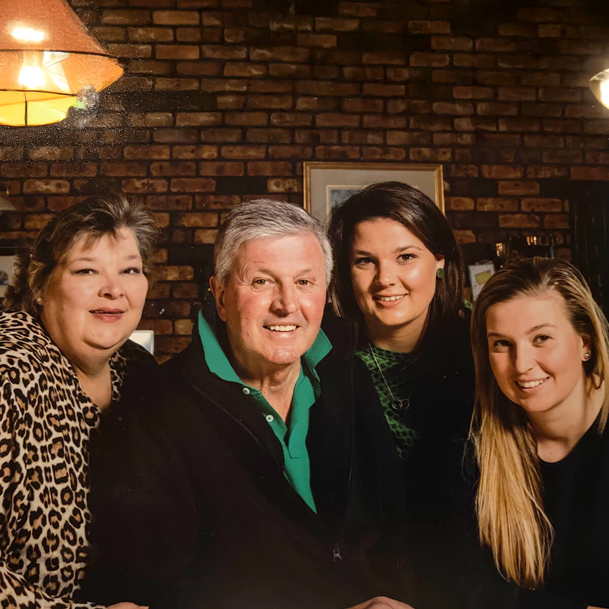 A smiling man sits in the center with three women beside him, against a brick wall and warm lighting.