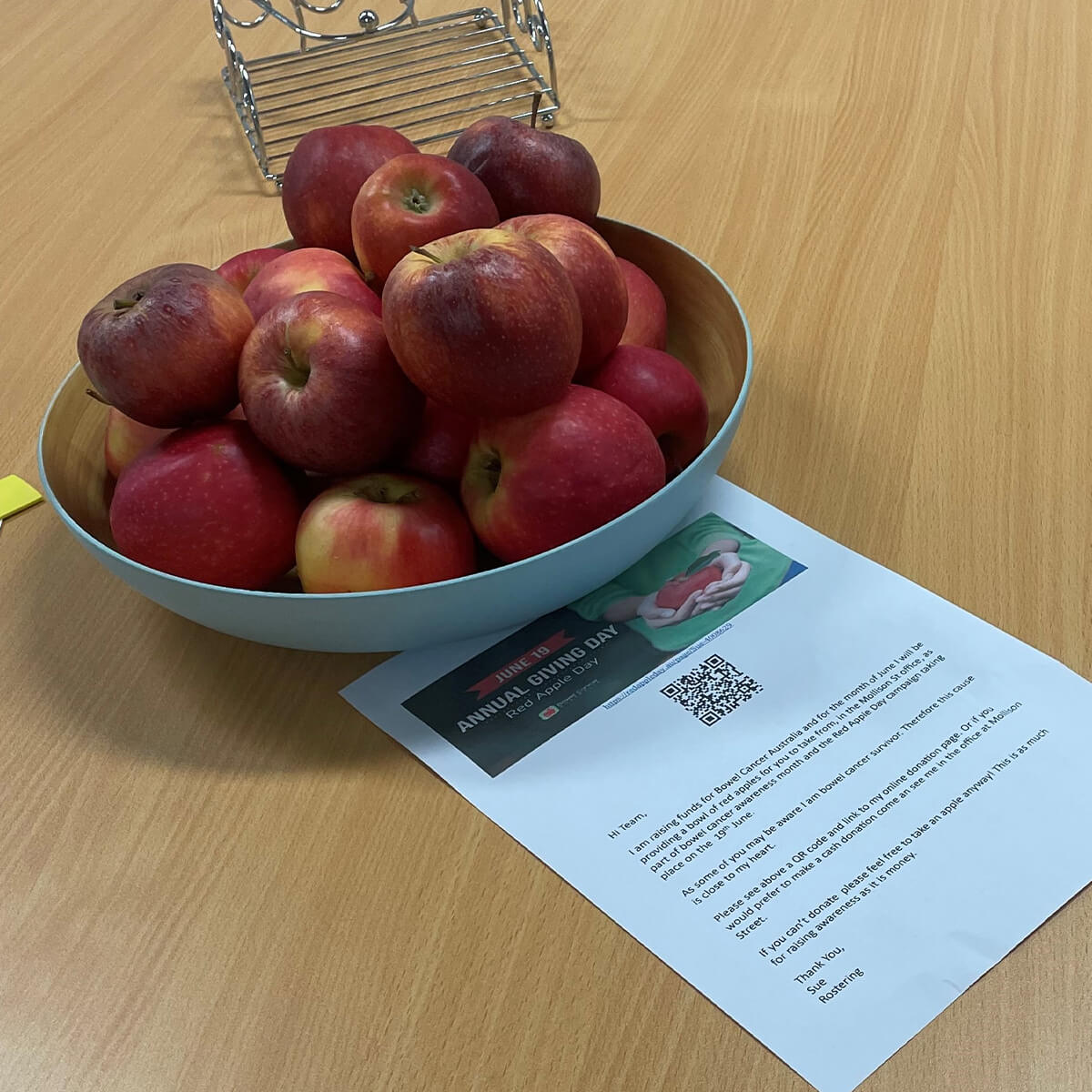 A bowl of red apples on a wooden table next to a printed flyer about Annual Giving Day.