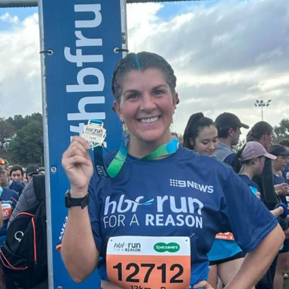 A smiling woman in a blue shirt holds a medal, standing in a crowd at a running event.
