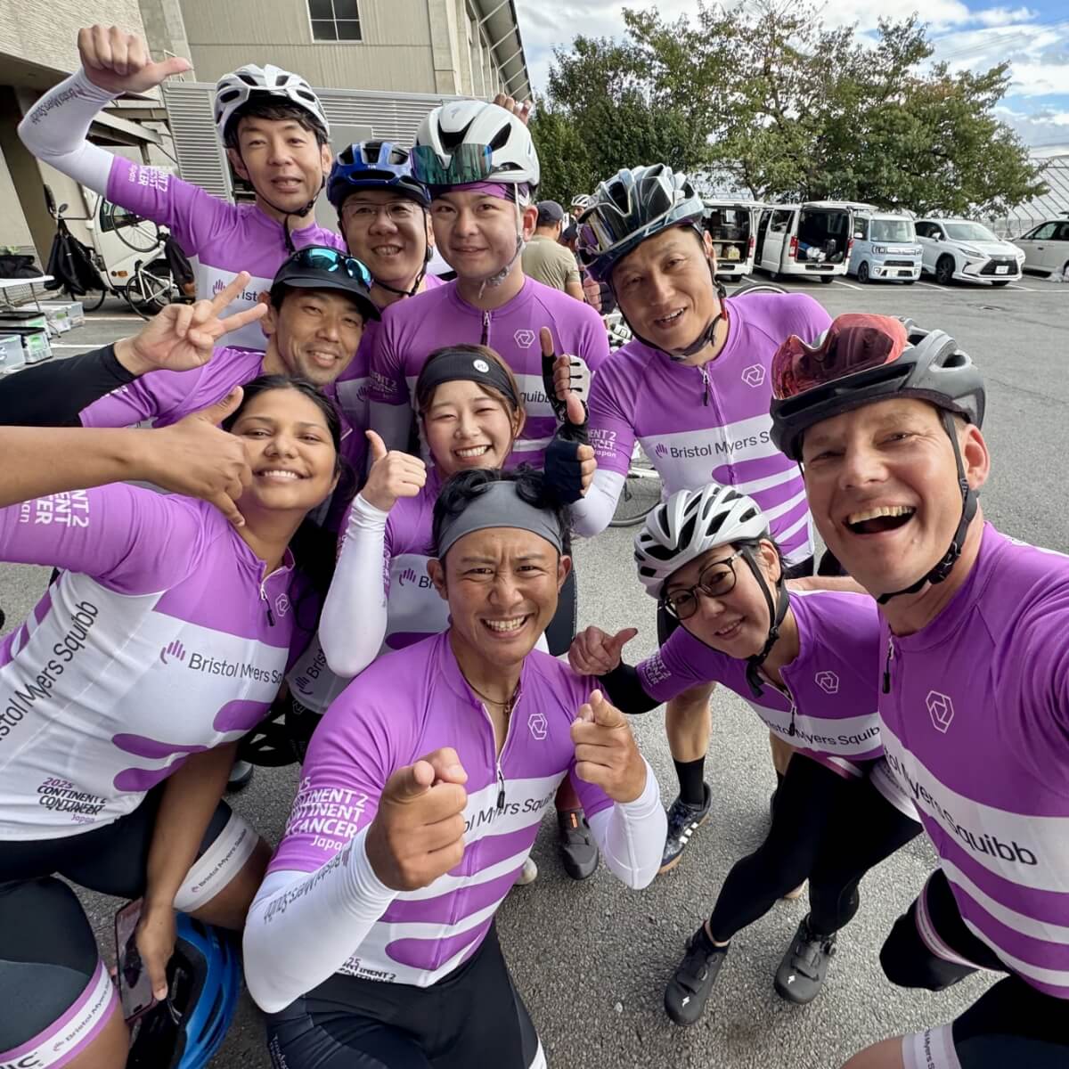 A group of cyclists in purple jerseys poses for a photo, smiling and giving thumbs up outdoors.