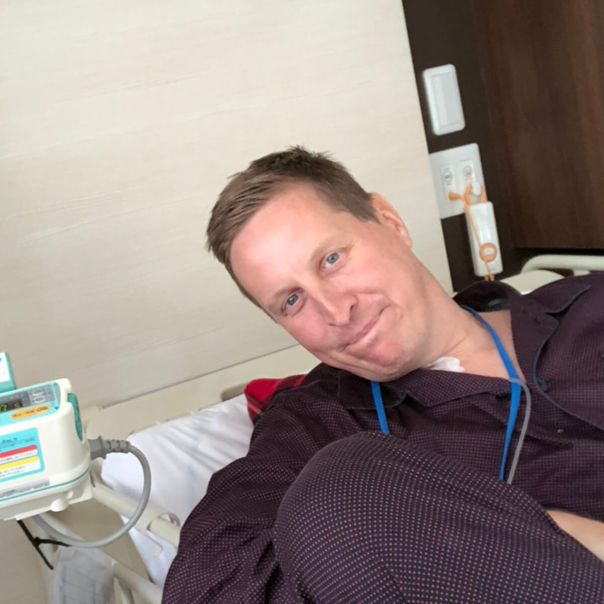 A man in a hospital bed smiles at the camera, wearing a dark shirt with a blue lanyard and patterned pants.
