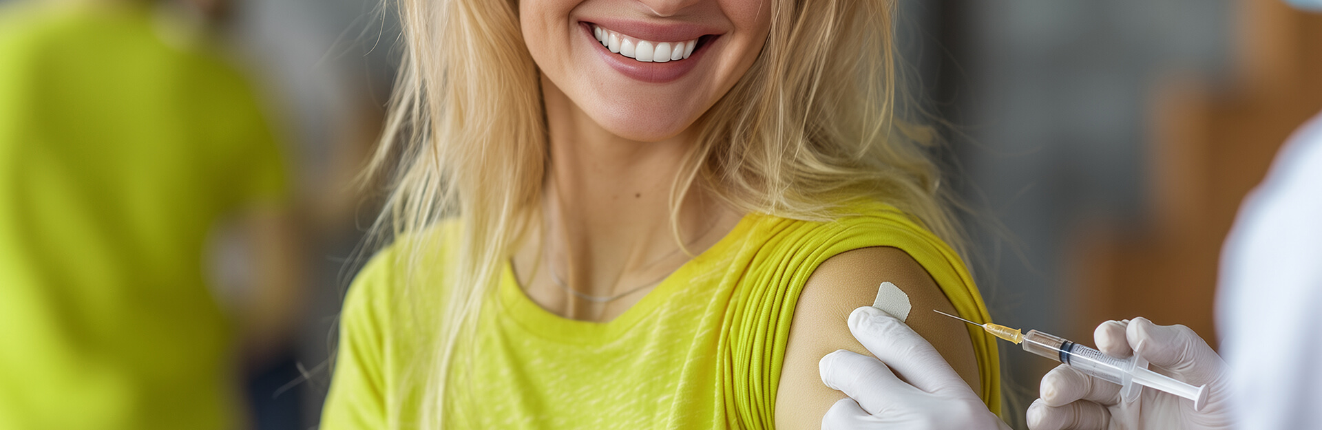 A smiling woman in a yellow shirt receives a vaccination in her upper arm from a healthcare professional.