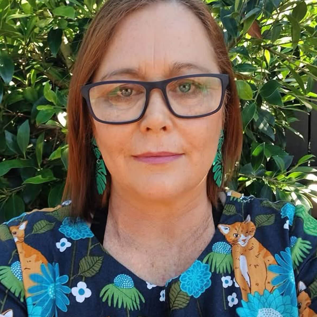 A woman with glasses and earrings stands in front of green foliage, wearing a floral-patterned top featuring a cat.