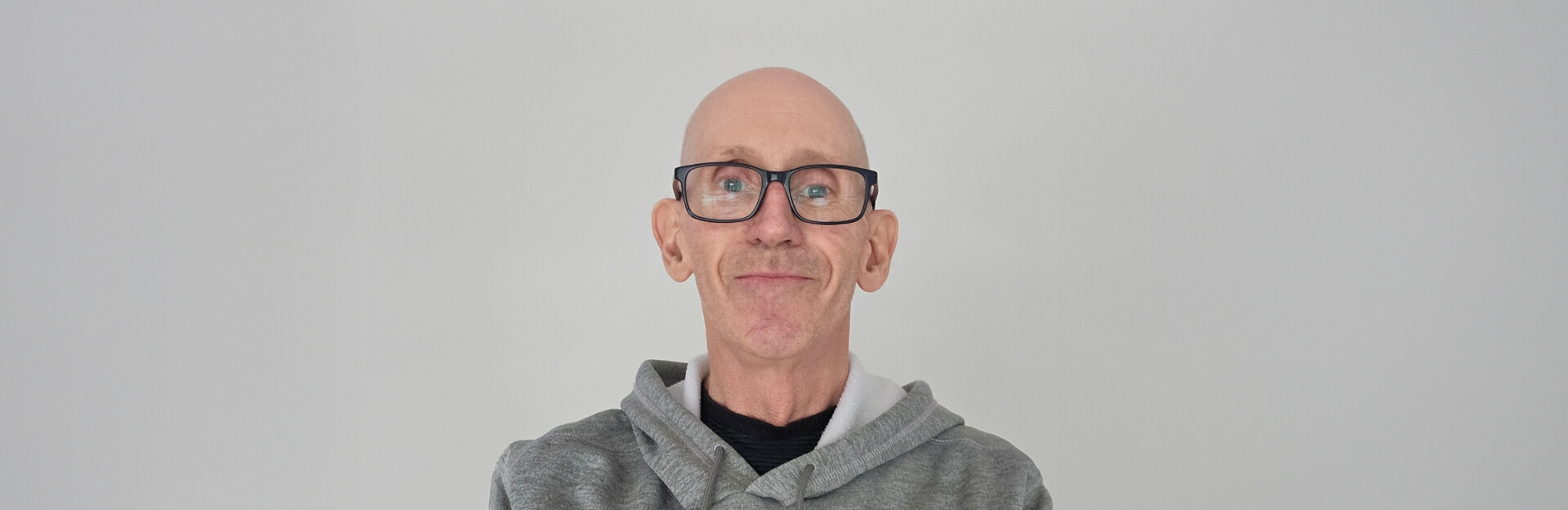A bald man in glasses smiles, wearing a gray hoodie against a plain light background.