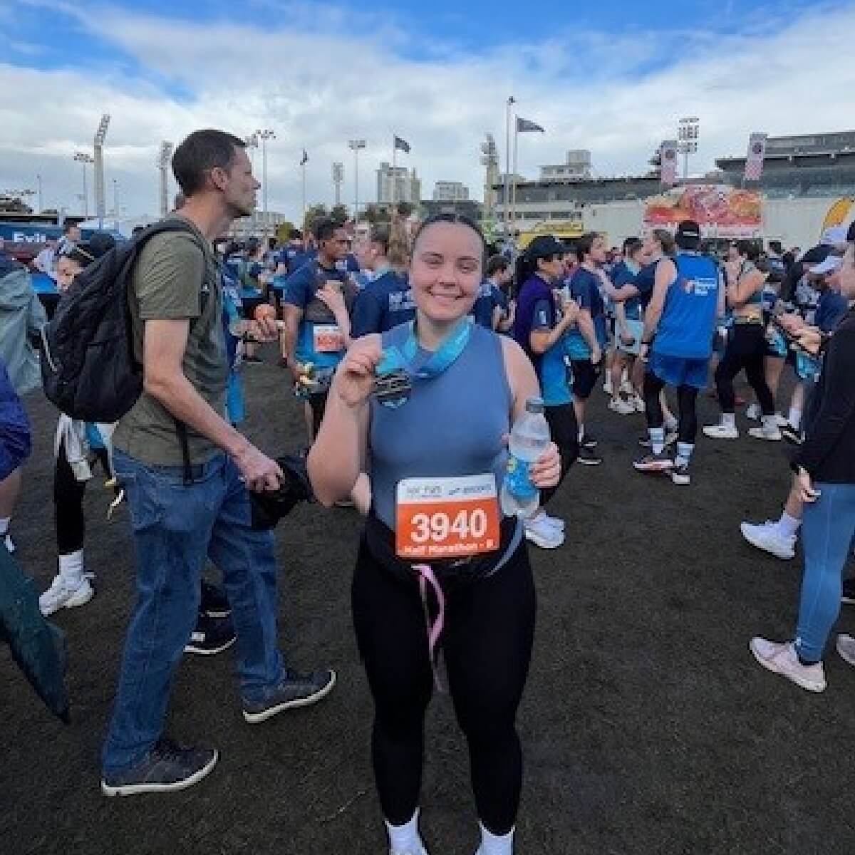 A smiling woman holds a medal and water bottle, surrounded by runners in a race setting.