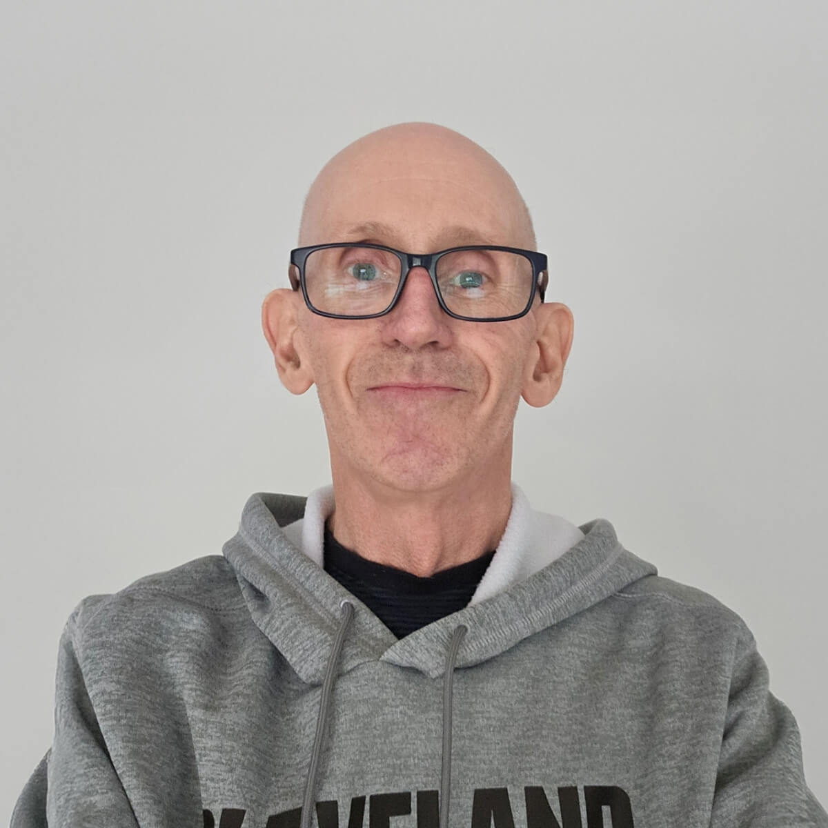 A bald man with glasses smiles, wearing a gray hoodie against a plain white background.