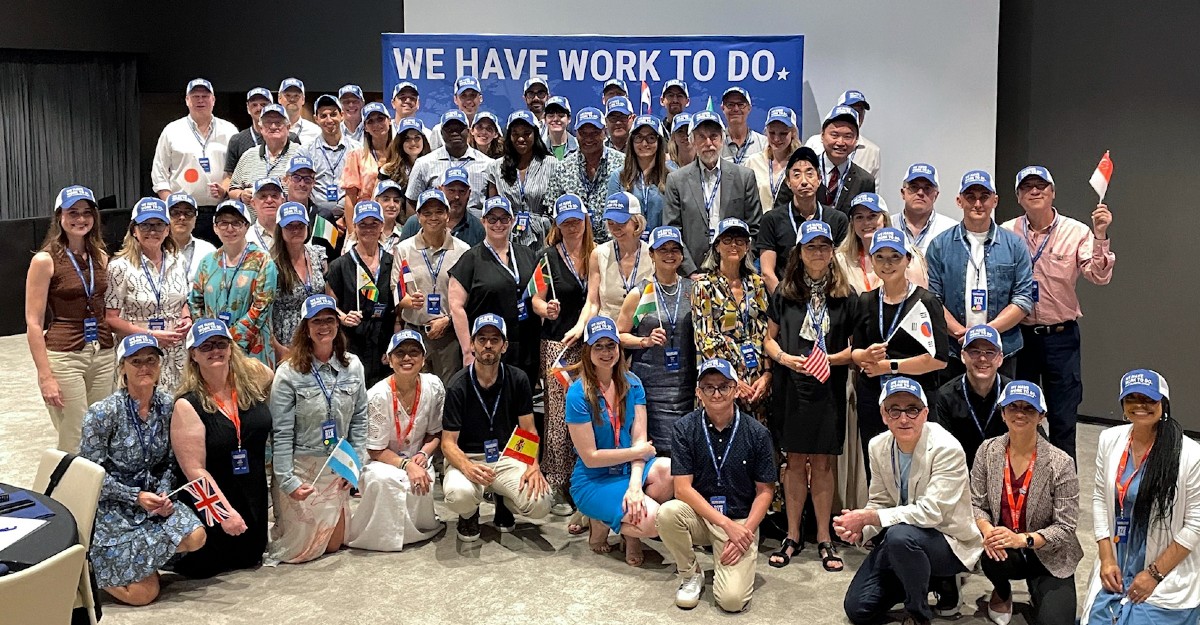 A diverse group of people wearing blue hats poses together in front of a banner that reads "WE HAVE WORK TO DO."