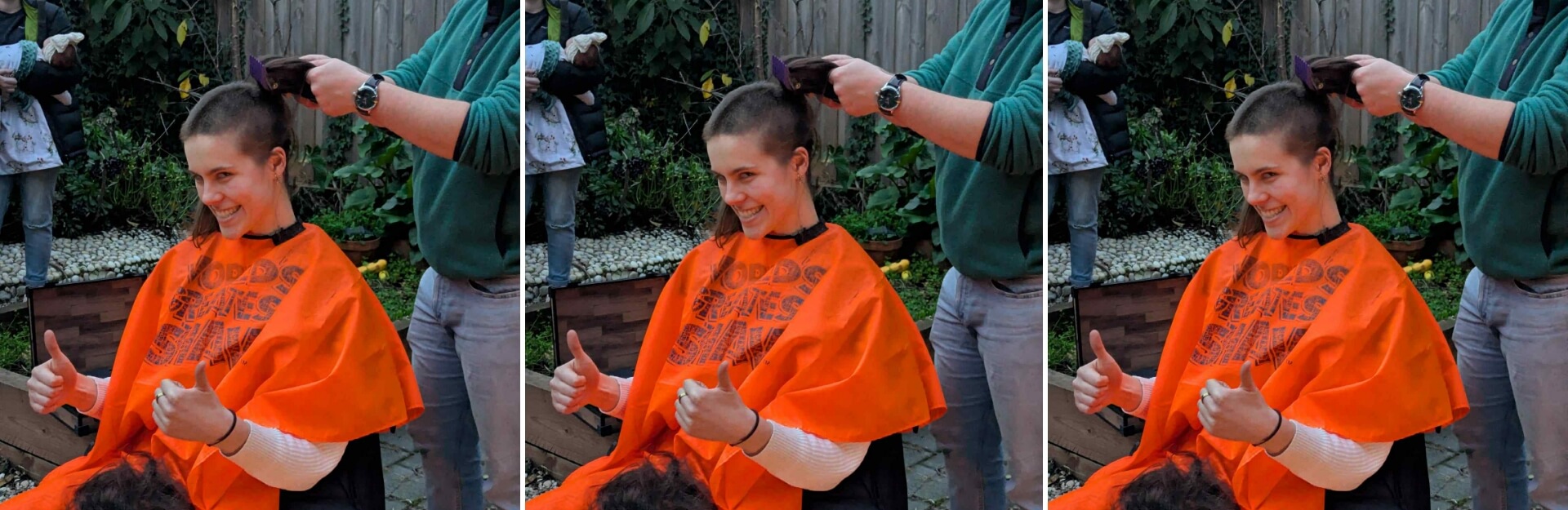 A person in an orange cape smiles and gives a thumbs up while getting their hair cut short outdoors.