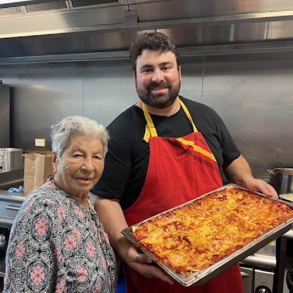 A smiling older woman stands next to a man in a red apron holding a tray of lasagna in a kitchen setting.