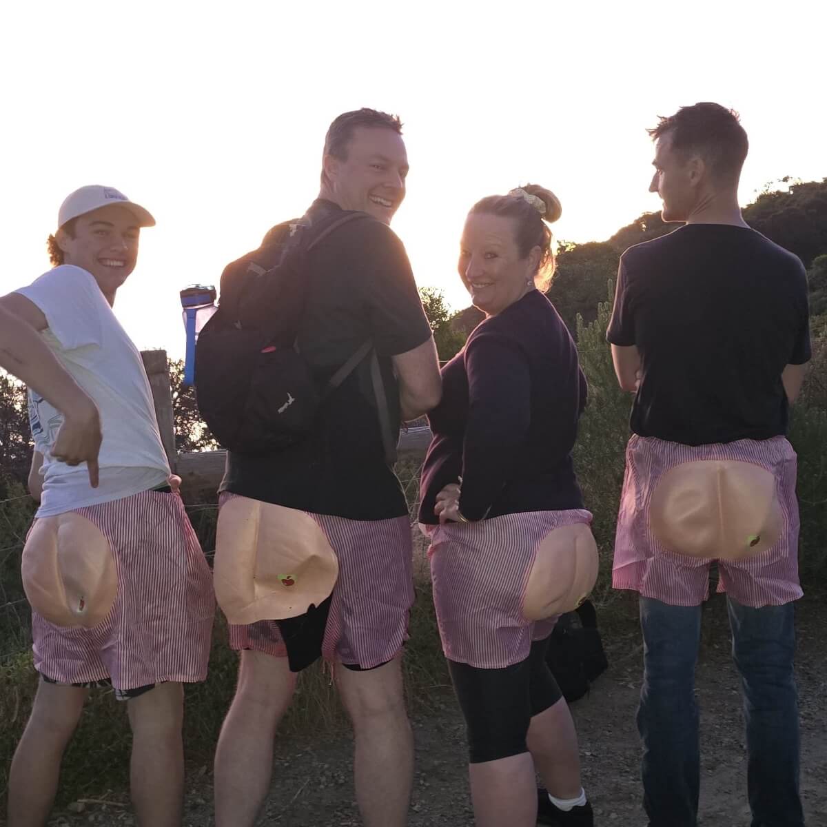 Four people stand outdoors, wearing shorts with fake buttocks, smiling at the camera during sunset.