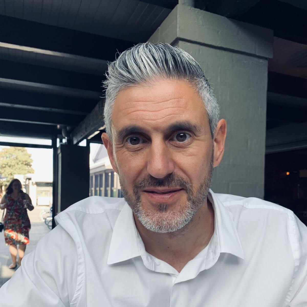 A man with short gray hair and a beard is smiling, wearing a white shirt, with a woman in a floral dress in the background.