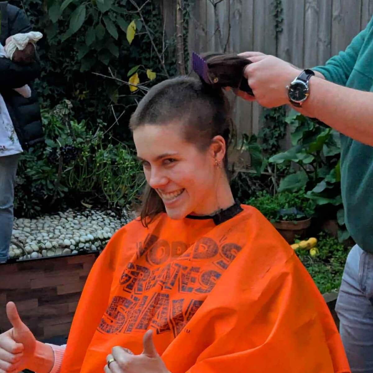 A woman in an orange cape smiles and gives thumbs up while someone cuts her hair in a garden setting.