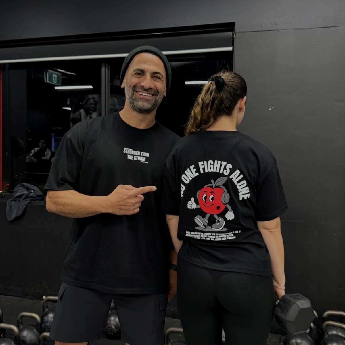 A smiling man points at a woman wearing a black shirt with a cartoon cherry and the phrase "NO ONE FIGHTS ALONE."