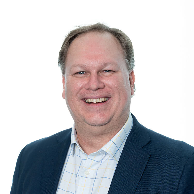 A smiling man with short hair wearing a blue blazer over a checkered shirt, set against a white background.