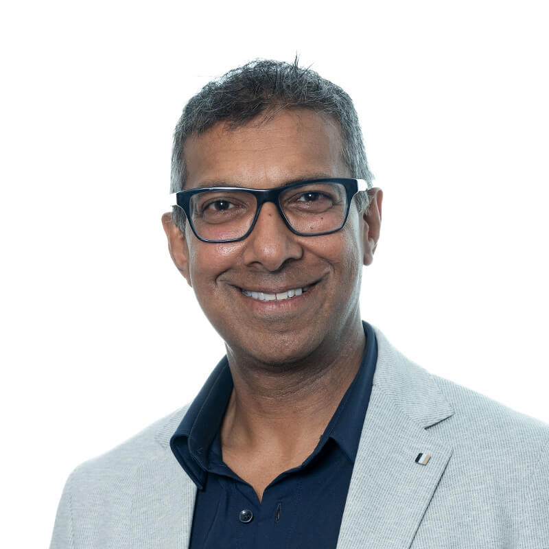 A smiling man with glasses, wearing a light gray blazer over a dark shirt, against a white background.