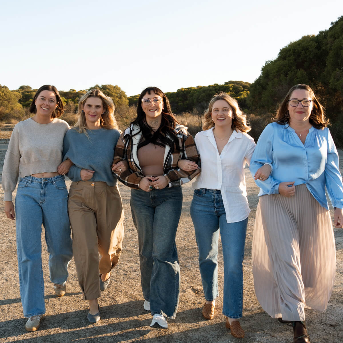 Five women walk together outdoors, smiling, wearing casual outfits, with trees in the background and a clear sky.