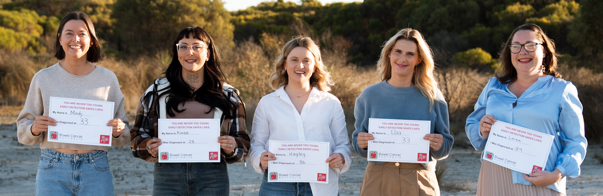Five women stand outdoors holding signs with their names and ages at diagnosis for bowel cancer awareness.
