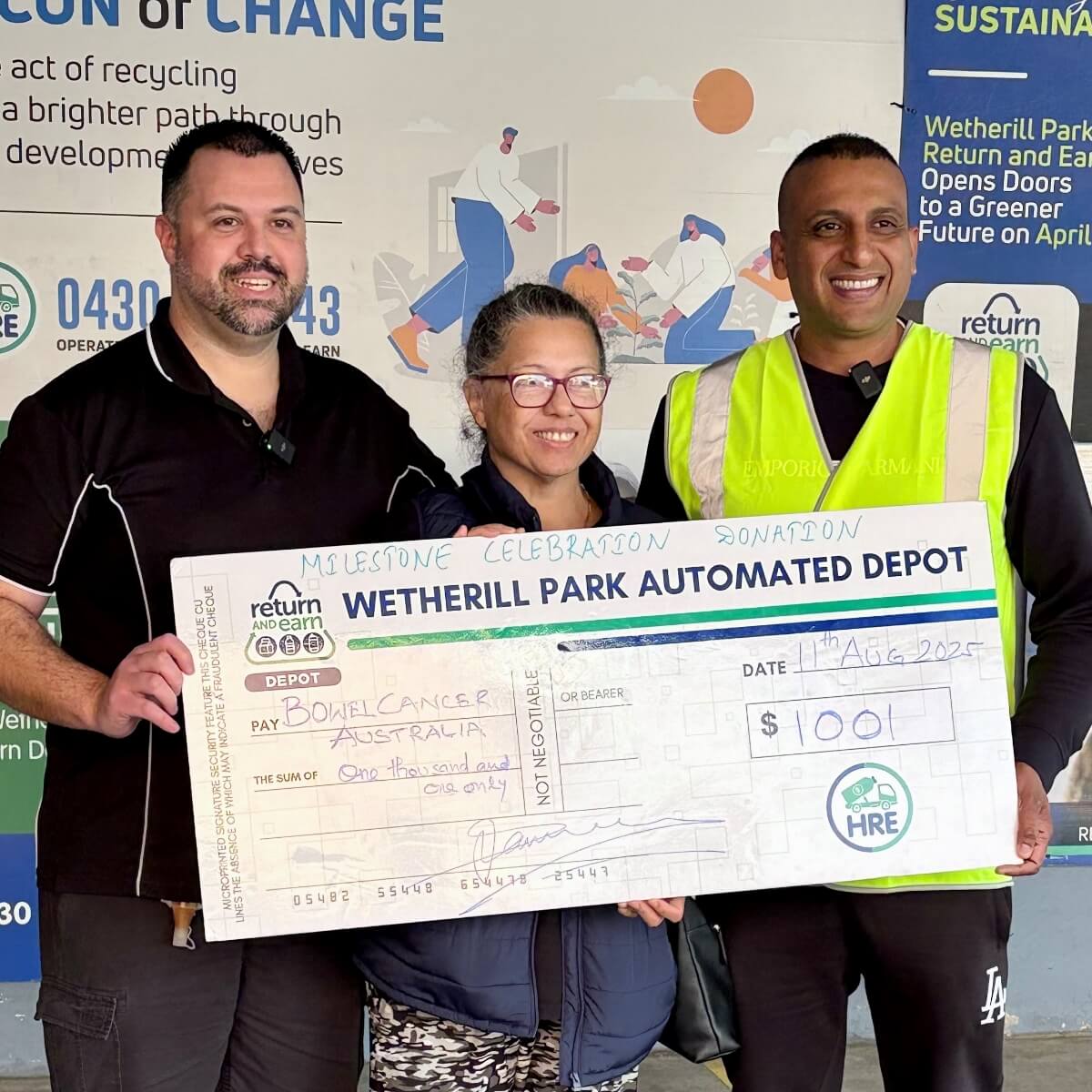 Three individuals hold a large donation check for Bowel Cancer Australia, in front of a recycling depot.