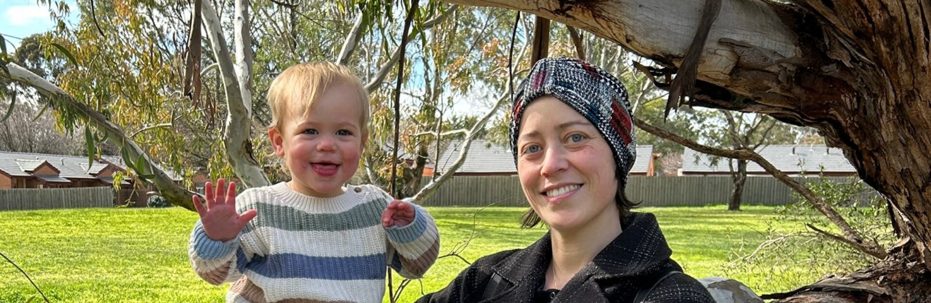 A woman and a toddler smile in a park, surrounded by trees and green grass.