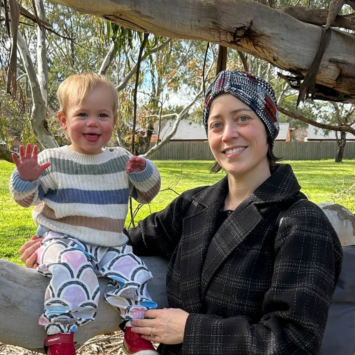 A woman and a toddler are sitting on a tree branch in a green park. The toddler is smiling and waving.