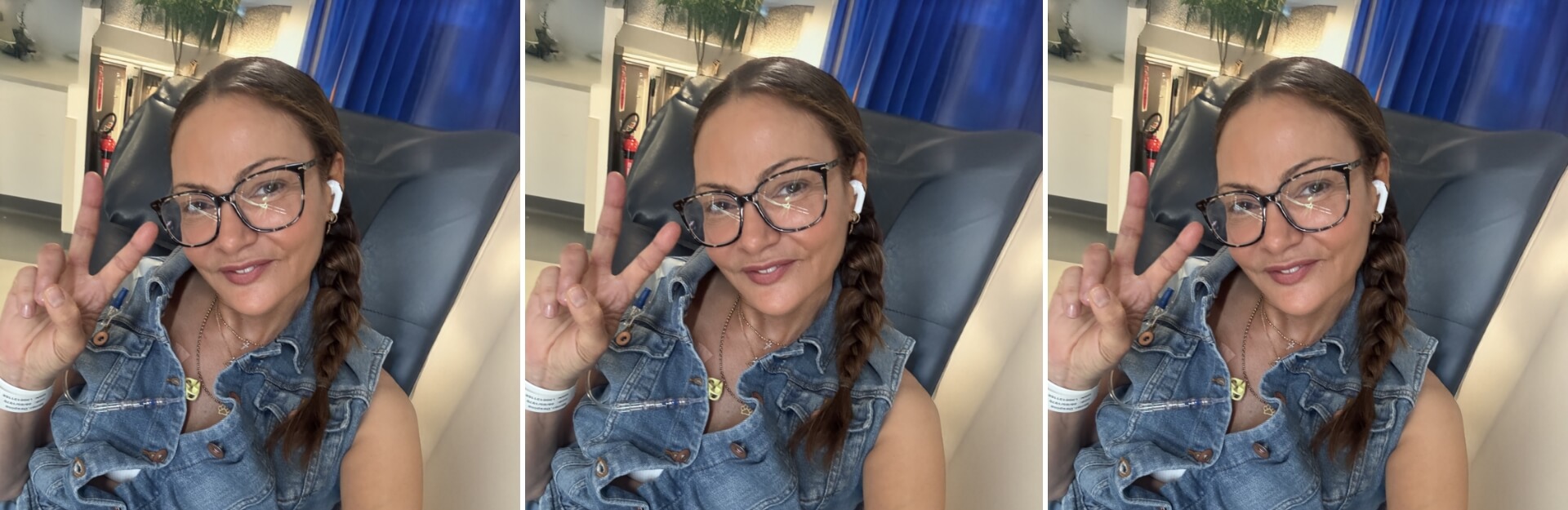 A woman with glasses and earbuds smiles and makes a peace sign, sitting in a chair with a blue curtain backdrop.