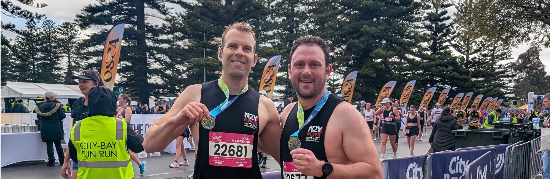 Two male runners pose with medals after completing a marathon, with trees and event banners in the background.