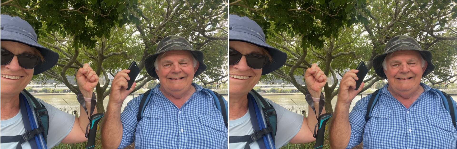 Two smiling individuals, one holding a phone, stand under a tree by a river, wearing hats and casual clothing.