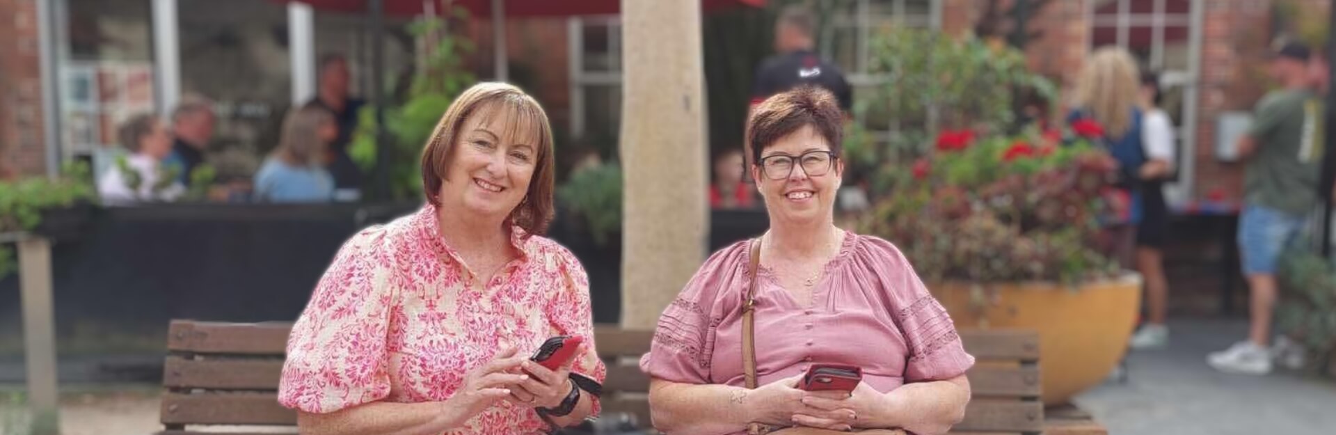 Two women sitting on a bench, smiling and holding smartphones, with a garden and people in the background.