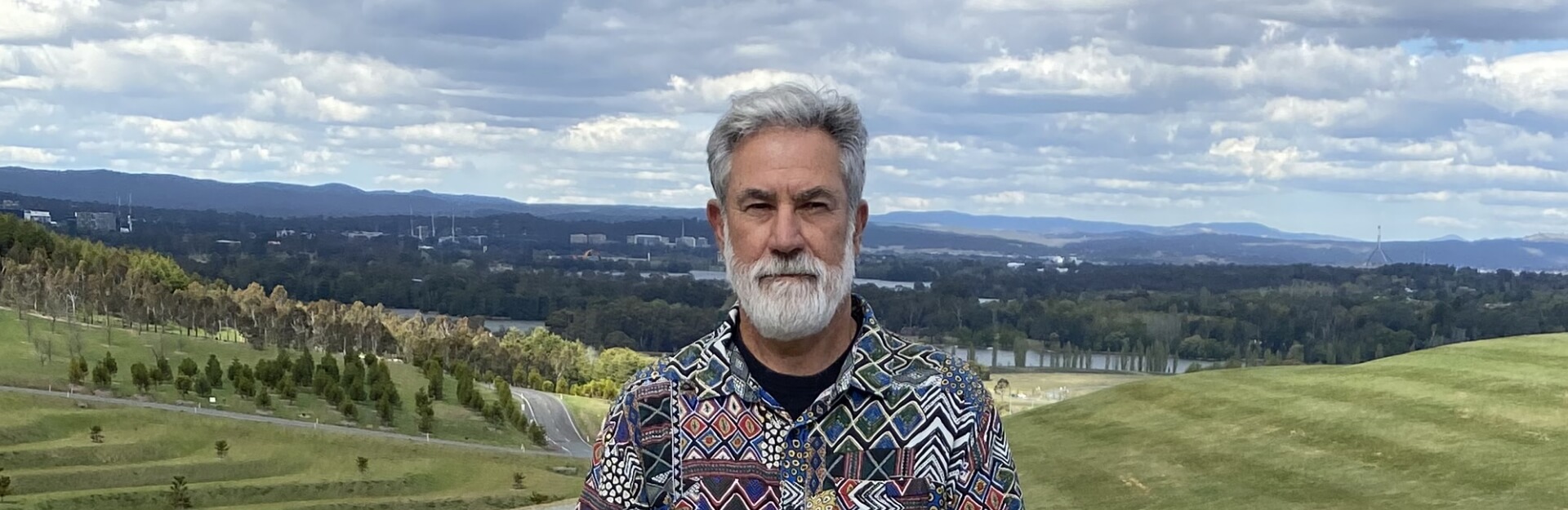 A man with a gray beard stands outdoors, wearing a colorful patterned shirt, with rolling hills and a cloudy sky in the background.