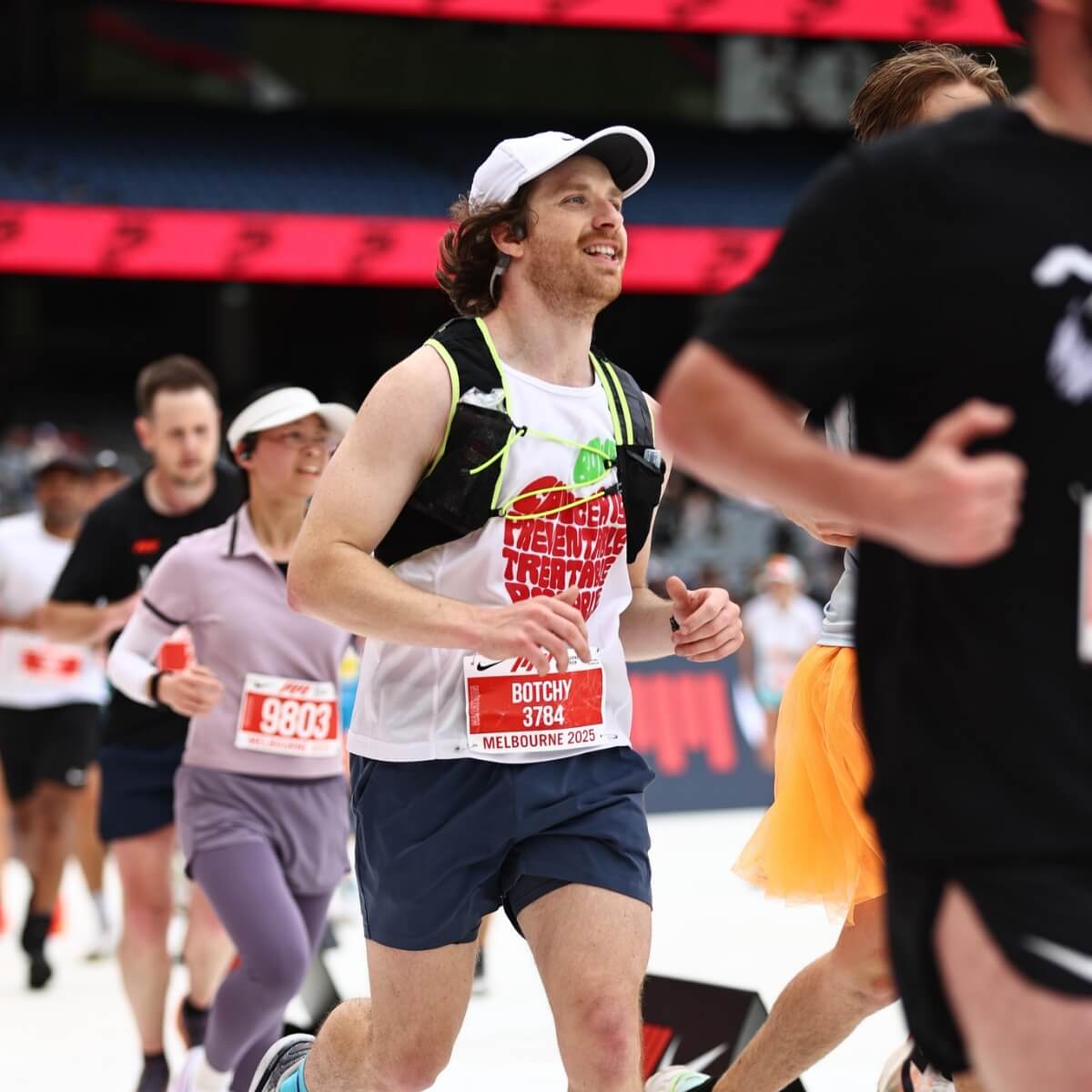 A group of runners in athletic gear participating in a race, with one man smiling and wearing a white tank top.