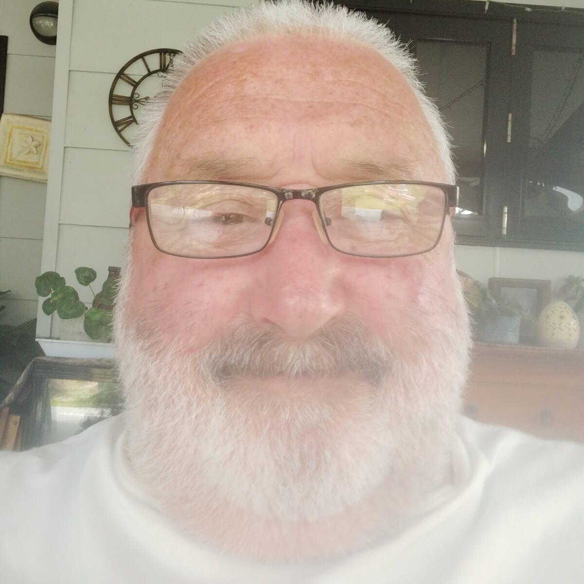 A smiling man with glasses and a beard, seated indoors with plants and decorative items in the background.