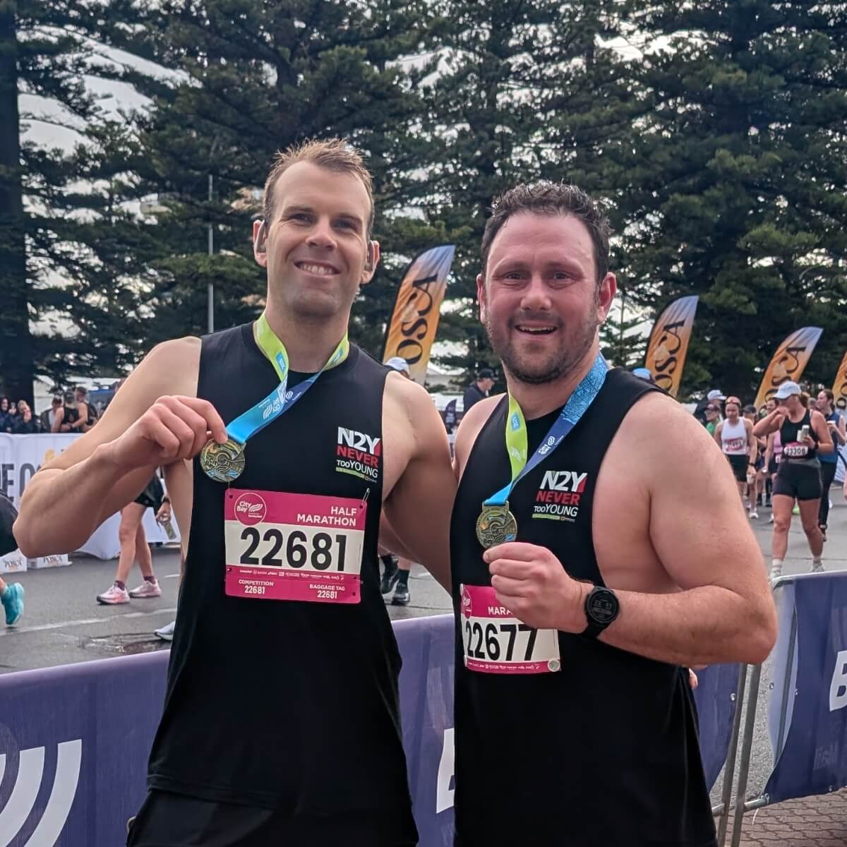 Two male runners in black tank tops hold medals, smiling at the camera with a race backdrop and participants behind them.