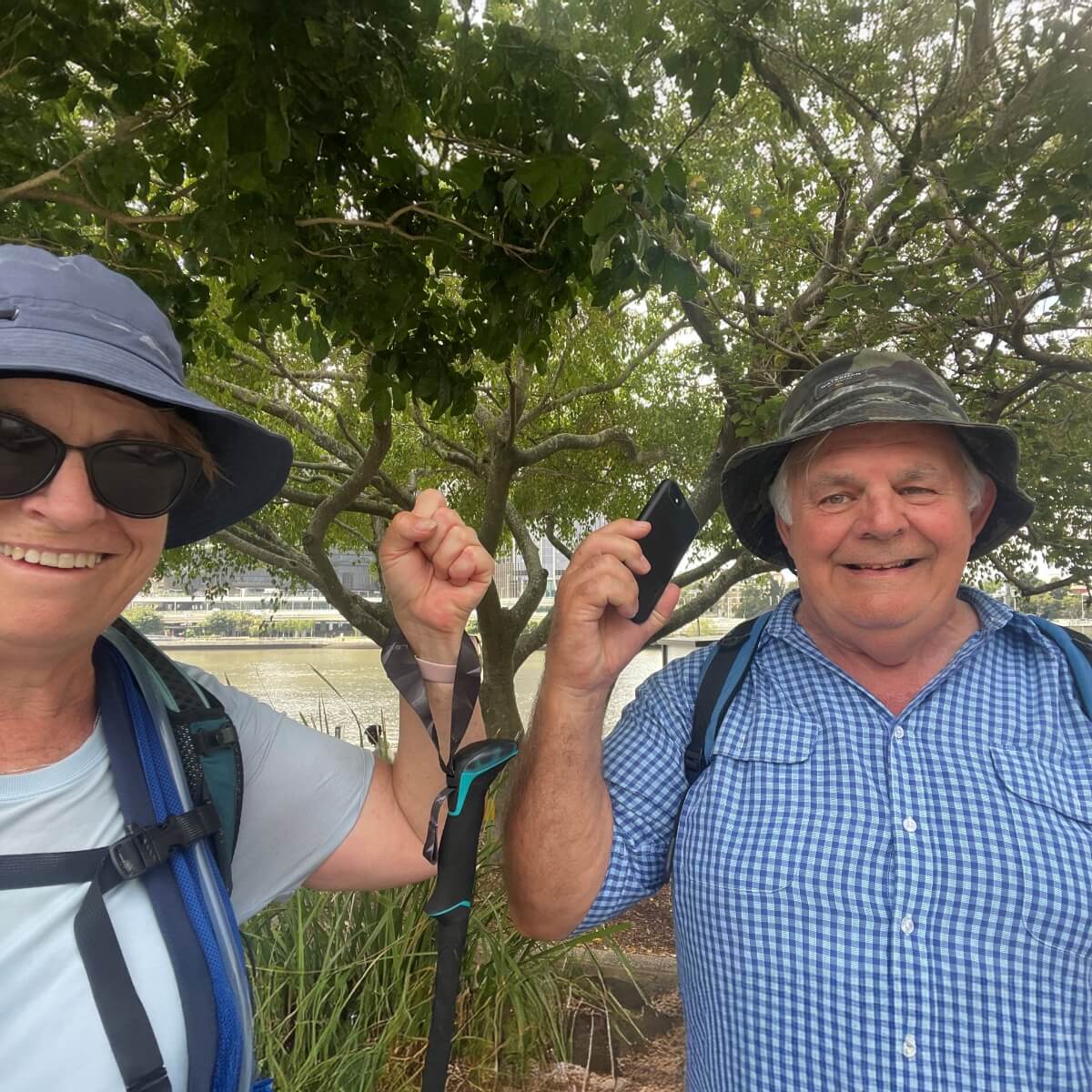 Two smiling individuals outdoors, one holding a phone and the other raising a fist, with greenery in the background.