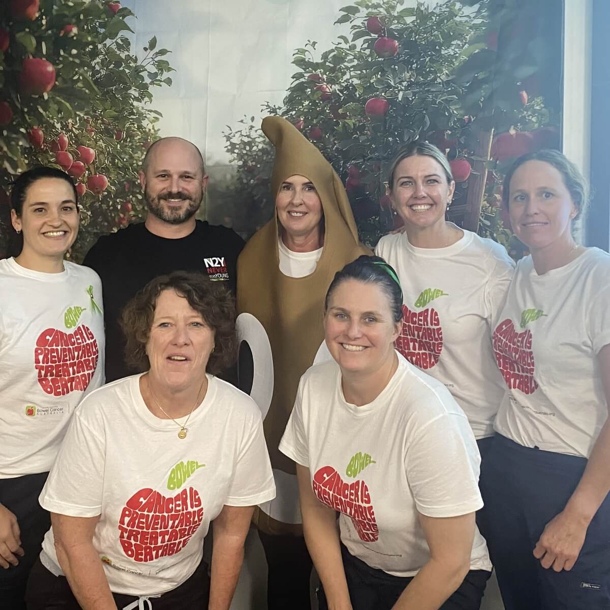 A group of seven people posing together, some wearing t-shirts with a cancer awareness message, in an apple orchard setting.
