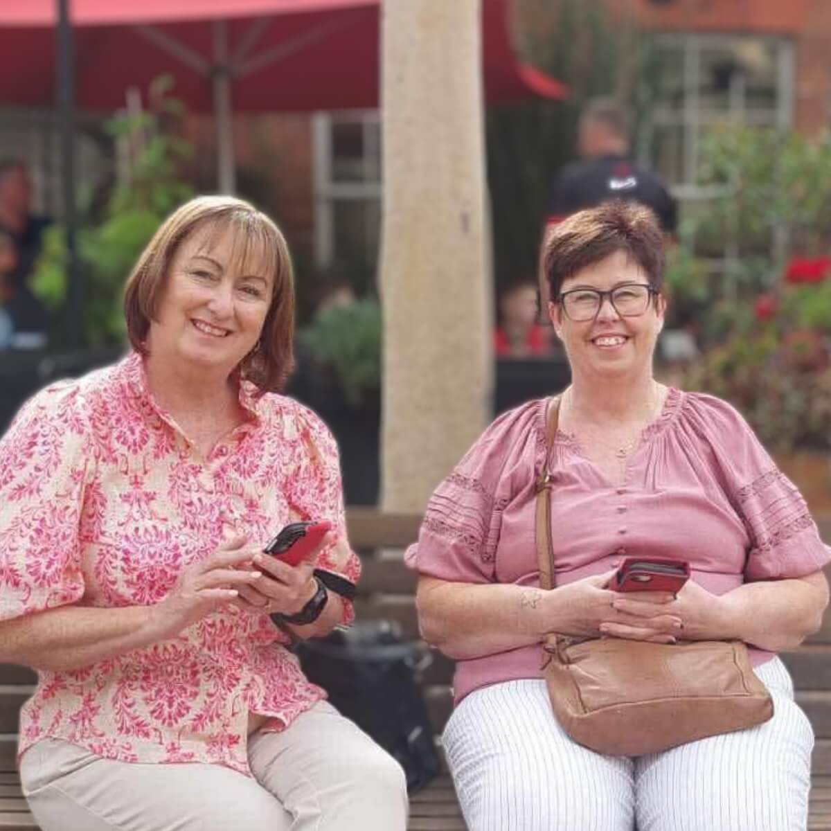 Two women sitting on a bench, smiling and holding smartphones, with greenery and people in the background.