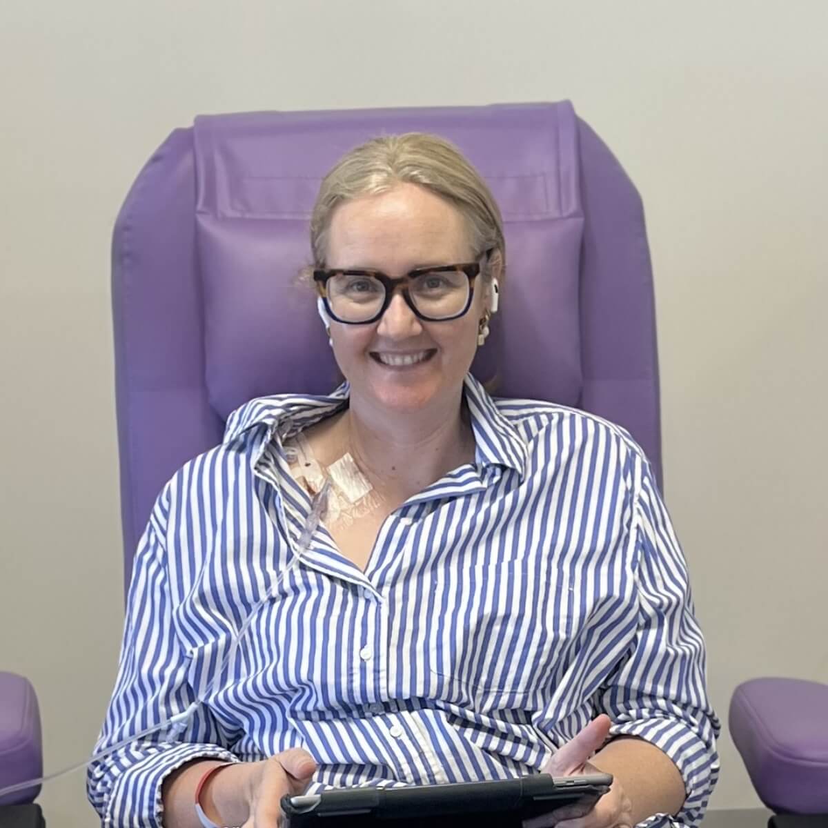 A smiling woman with glasses sits in a purple chair, wearing a striped shirt and earbuds, holding a tablet.