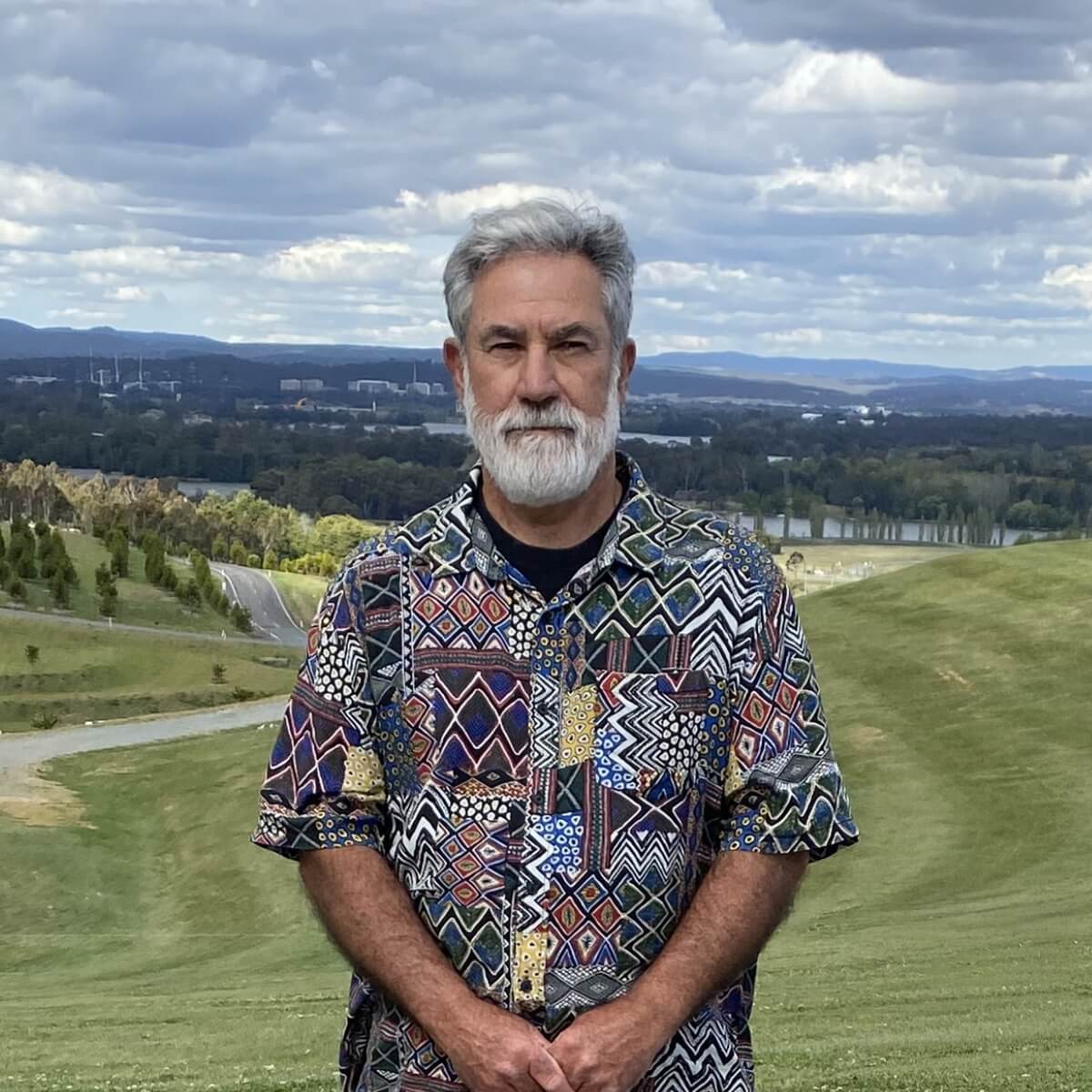 A man with a beard stands on a grassy hill, wearing a colorful patterned shirt, with a cloudy sky and distant landscape behind him.