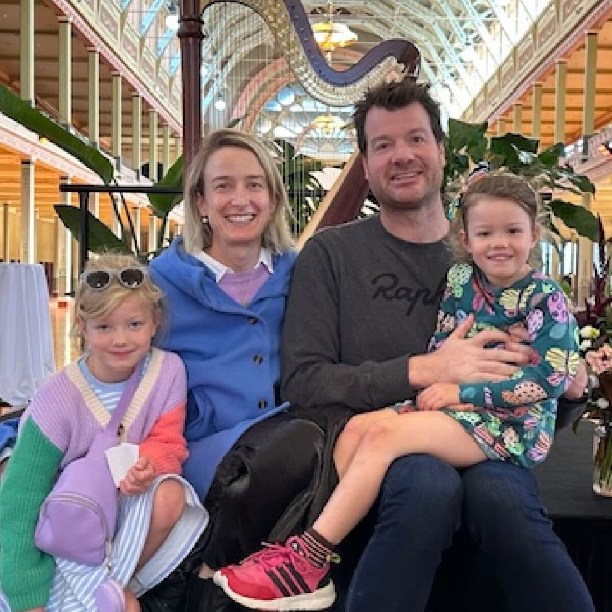 A family of four smiles together in a colorful indoor space with a harp and plants in the background.