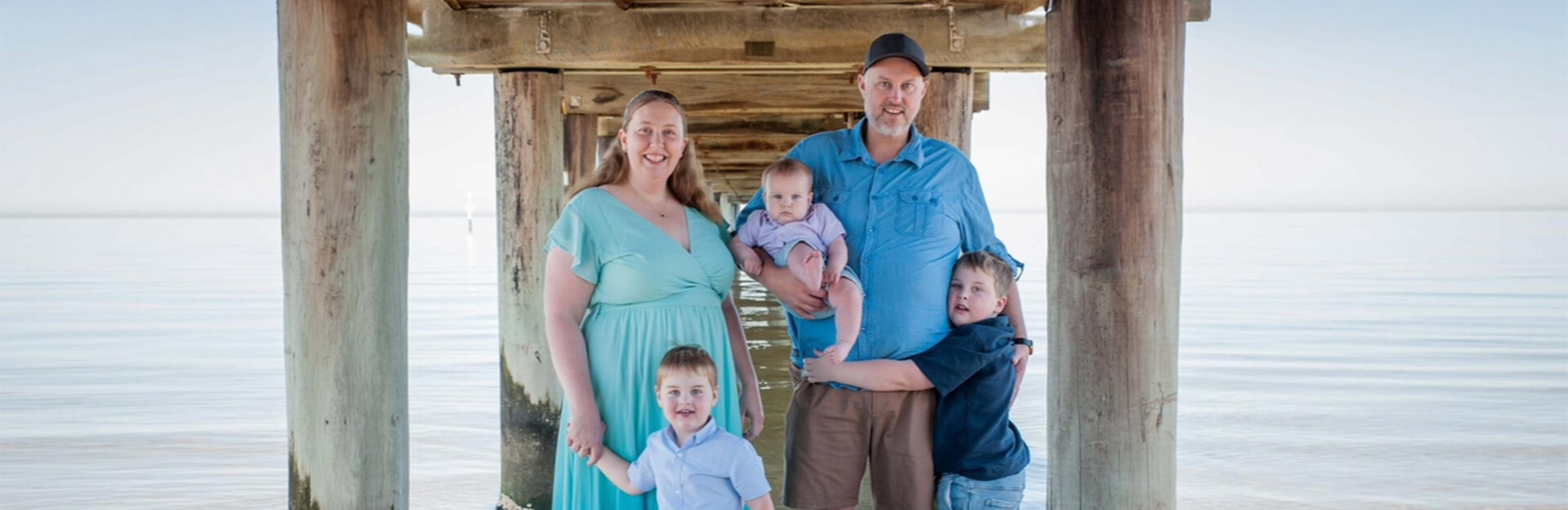 A family of five stands under a pier, with calm water in the background. They are smiling and dressed casually.
