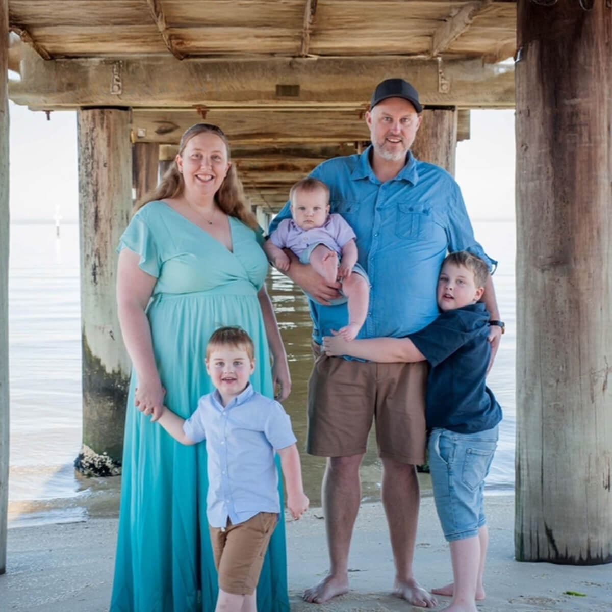A family of four stands under a pier, smiling. The mother holds hands with a young boy, while the father holds a baby.