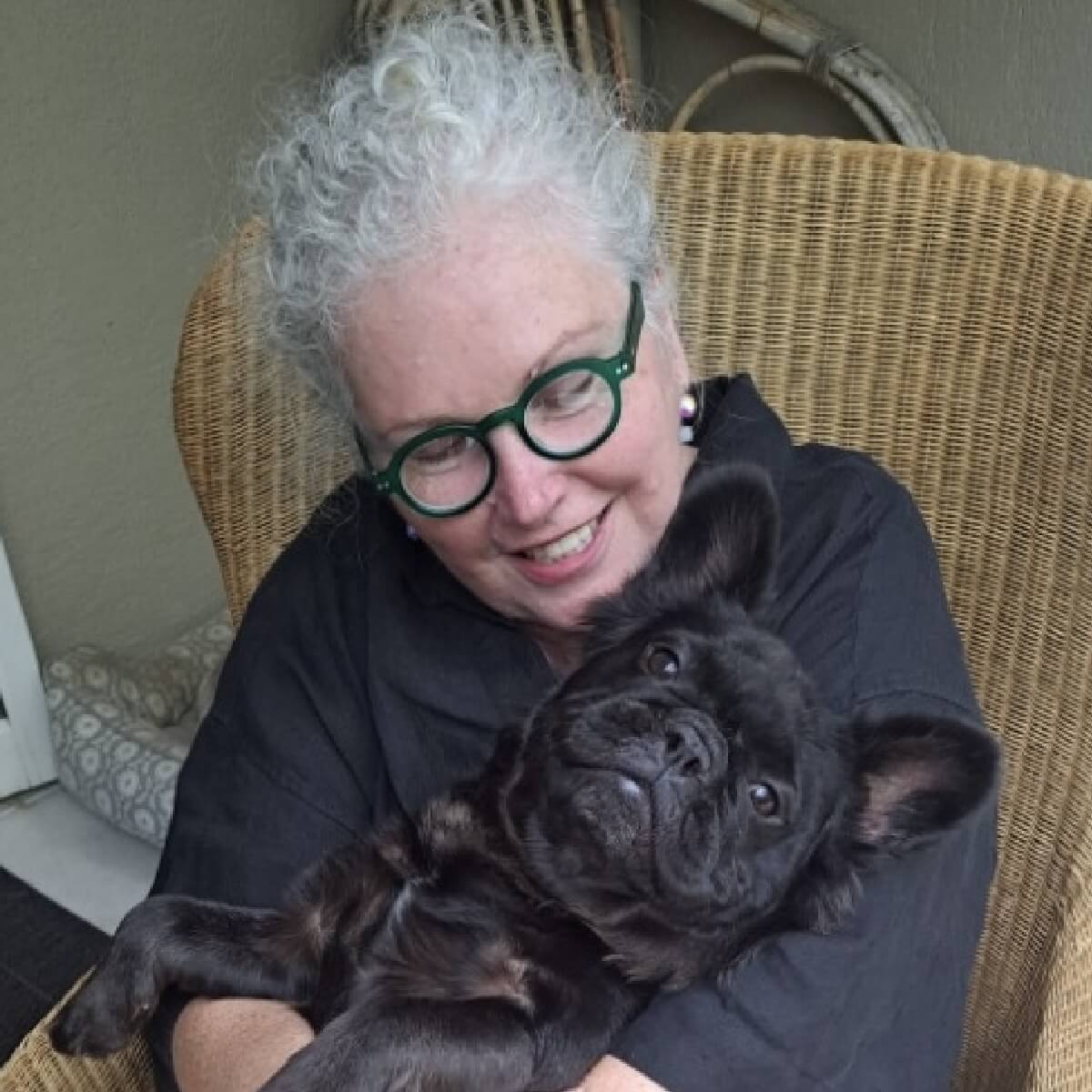 A woman with curly gray hair and green glasses smiles while holding a black French Bulldog in her lap.