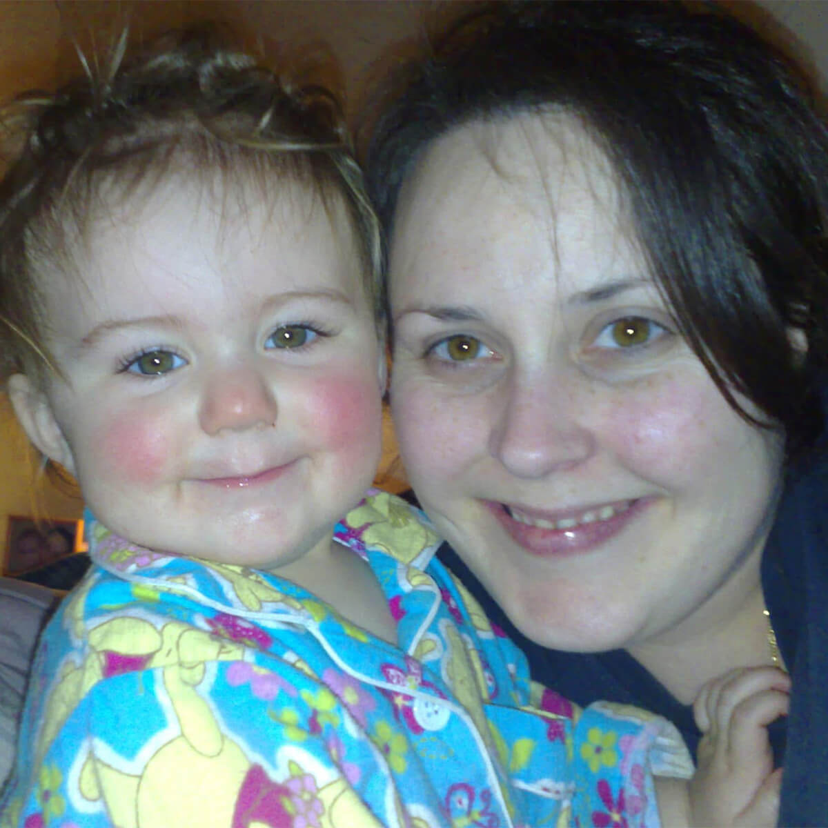 A smiling woman and toddler pose closely together, both wearing colorful pajamas. They have joyful expressions.