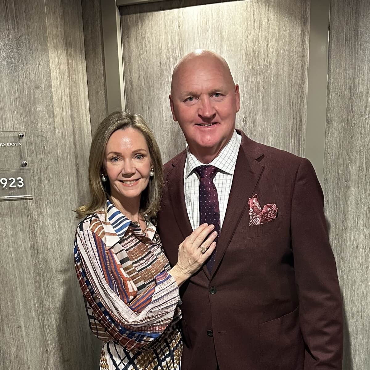 A woman in a patterned blouse poses with a man in a burgundy suit by a door marked "923."