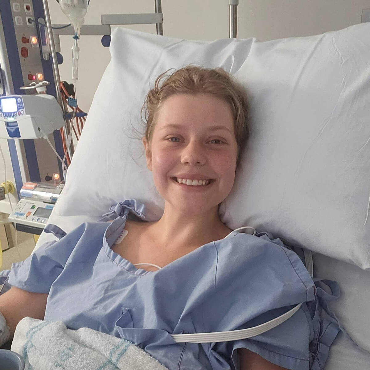 A smiling young woman in a hospital bed, wearing a blue gown, with medical equipment visible in the background.