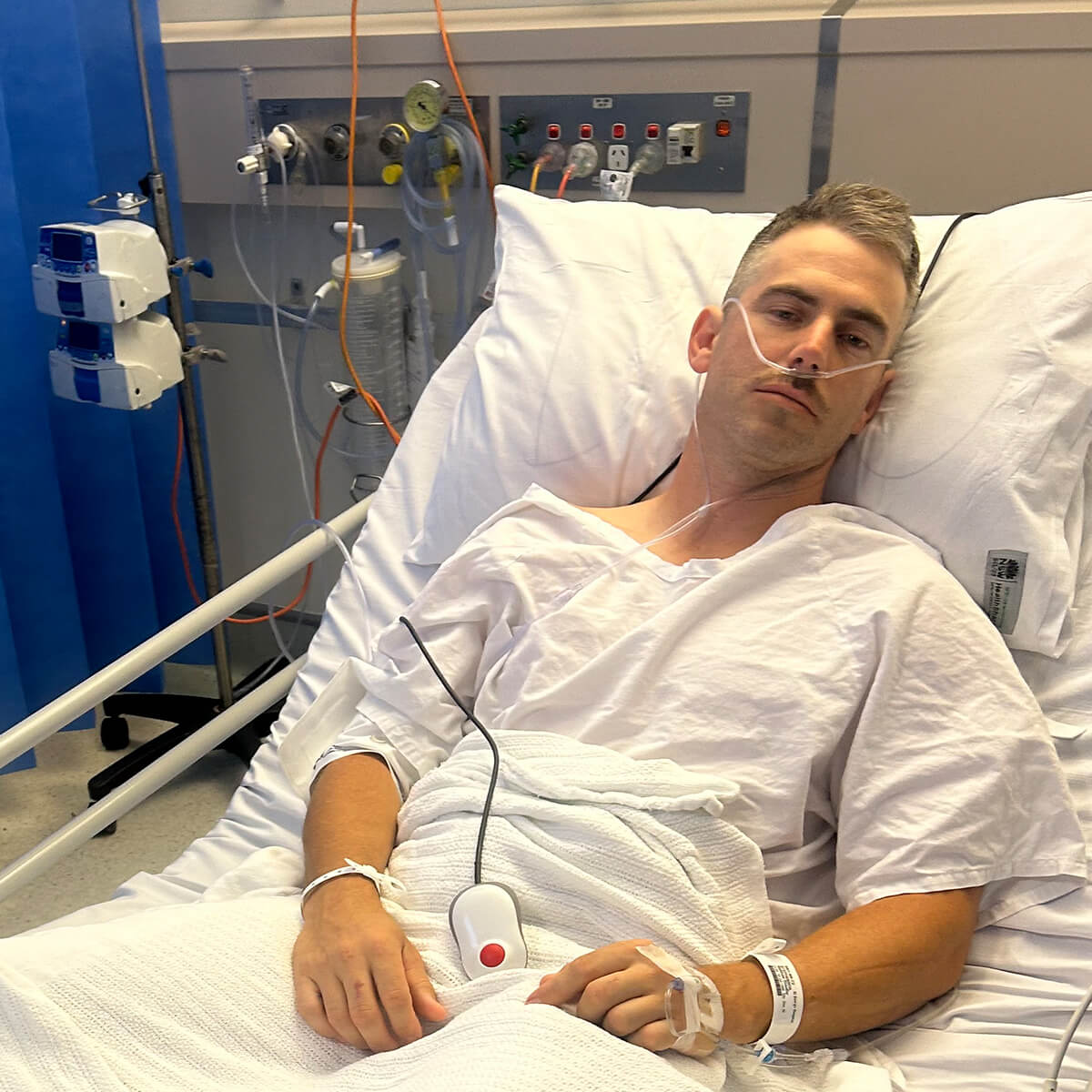 A patient in a hospital bed wearing a gown, with medical equipment and oxygen tubes visible around them.
